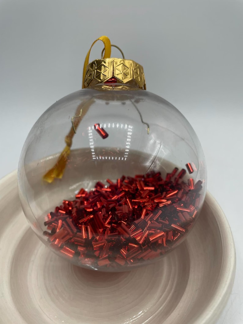 May include: A clear, spherical glass ornament filled with small, metallic red cylindrical beads. The ornament features a gold-colored metal cap with a geometric star pattern and a yellow ribbon loop for hanging.