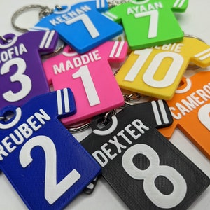 May include: A collection of colourful, jersey-shaped keychains made of flexible material. Each keychain features a unique name and number in white, including names like Keenan, Ayaan, Sofia, Maddie, Albie, Reuben, Dexter, and Cameron with numbers.