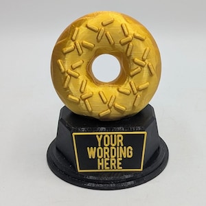 May include: A gold-coloured donut trophy with a black base. The base has a black plaque with the text "YOUR WORDING HERE".
