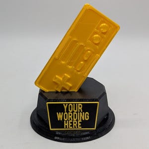 May include: A yellow 3D printed trophy shaped like a retro video game controller. The base is black with a black plaque that says "YOUR WORDING HERE".