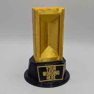 May include: A gold-colored trophy with a rectangular top and a black base. The trophy has the text "YOUR WORDING HERE" on a gold-bordered black plate. The trophy is likely a custom award.