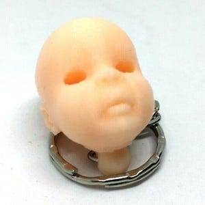 May include: A small, light pink baby doll head keychain with a silver metal key ring.