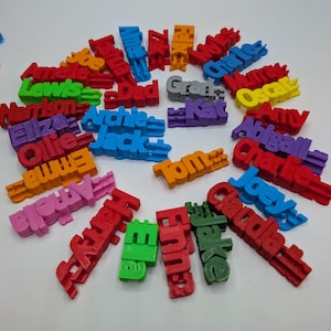 May include: A selection of brightly coloured 3D-printed name tags. Each tag displays a different name, such as "Jack", "Amy", and "Oscar", in a sans-serif font. The tags are in various colours, including red, blue, green, and yellow.