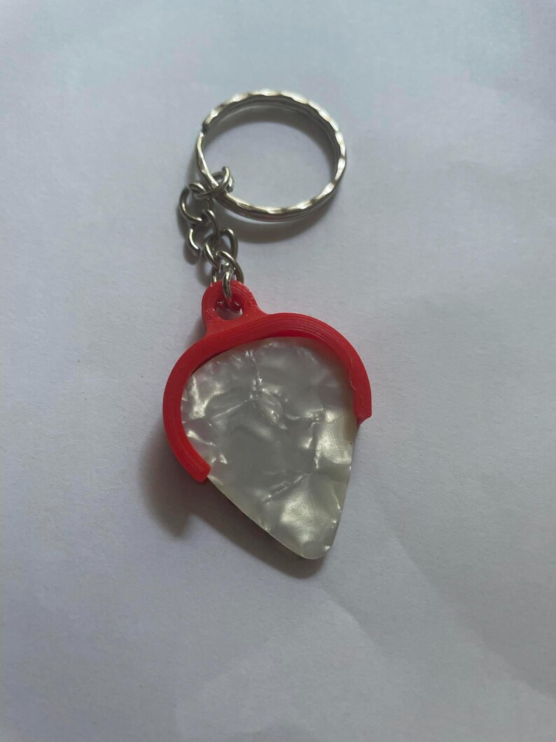 May include: A white guitar pick with a red plastic holder attached to a silver key chain.