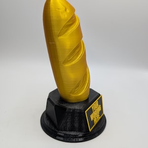 May include: A 3D-printed trophy featuring a golden baguette on a black base. The base has a rectangular plaque with the text "YOUR WORDING HERE" in black and yellow. The baguette has detailed ridges.