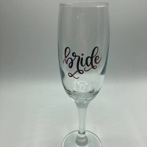 May include: A clear glass champagne flute with the word "bride" written in gold lettering.