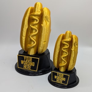 May include: Two gold-colored hot dog trophies with black bases. The hot dogs have a wavy mustard design. Each base has a gold-bordered black plaque with the text "YOUR WORDING HERE".