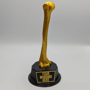 May include: A gold-colored bone-shaped trophy with a black base. The base has a gold-lettered plaque that reads "YOUR WORDING HERE". The trophy is likely a custom award or decorative item.