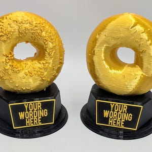 May include: Two gold bagel-shaped trophies on black bases. One bagel has a textured surface, the other is smooth. Each trophy has a black plaque with the text "YOUR WORDING HERE" in gold lettering. The trophies are on a white background.