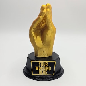May include: A gold-colored 3D printed hand sculpture in a pinched gesture, mounted on a black octagonal base. The base features a rectangular plaque with the text "YOUR WORDING HERE" in raised gold lettering against a black background.
