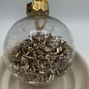 May include: A clear spherical glass ornament filled with numerous small, metallic gold-toned cylindrical beads. The ornament features a gold-colored metal cap with a geometric snowflake-like pattern and a yellow ribbon loop for hanging.