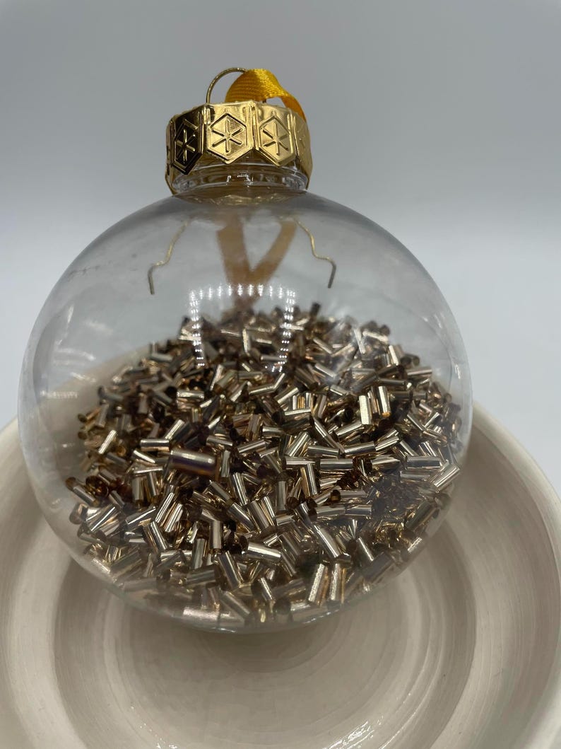 May include: A clear spherical glass ornament filled with numerous small, metallic gold-toned cylindrical beads. The ornament features a gold-colored metal cap with a geometric snowflake-like pattern and a yellow ribbon loop for hanging.