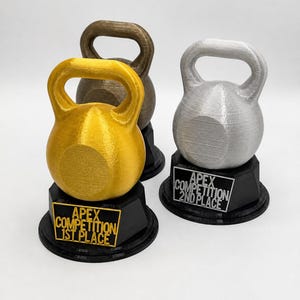 May include: Three kettlebell-shaped trophies on black circular bases. The front left trophy is gold with a plaque reading APEX COMPETITION 1ST PLACE. The front right trophy is silver with a plaque reading APEX COMPETITION 2ND PLACE. The back trophy is bronze.