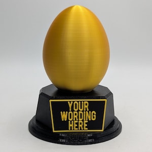 May include: A gold egg trophy with a black base. The base has a plaque with the text "YOUR WORDING HERE".