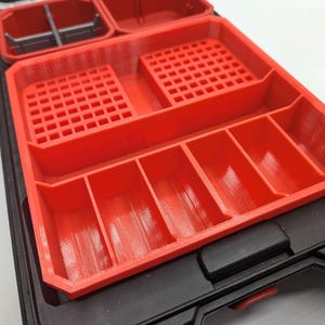 May include: A red plastic organiser with multiple compartments and a black base. The organiser has various sections, including slotted areas and divided compartments, designed for storing small items. The design suggests it's for tools or small parts.
