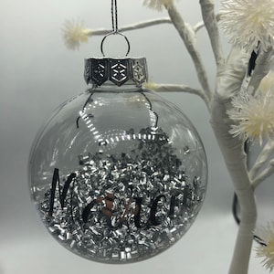 May include: A clear, spherical glass ornament filled with small, metallic silver rectangular beads. The exterior features the black cursive text "Marci" and a silver-toned metal cap with a snowflake pattern, suspended by a thin silver cord.