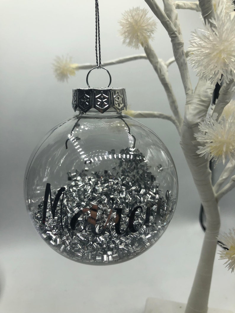 May include: A clear, spherical glass ornament filled with small, metallic silver rectangular beads. The exterior features the black cursive text "Marci" and a silver-toned metal cap with a snowflake pattern, suspended by a thin silver cord.