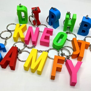 May include: A selection of brightly coloured 3D-printed keyrings, each in the shape of a letter. The keyrings are in various colours, including blue, red, yellow, green, pink, and orange, and are attached to silver key rings.