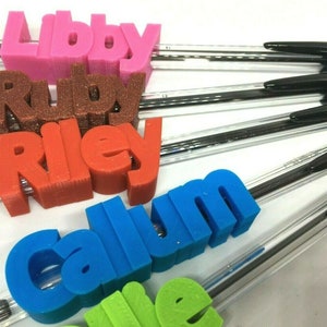 May include: A set of six personalized pens with names printed in 3D on the top of each pen. The names are Libby, Ruby, Riley, Callum, and two other names that are not visible in the image. The pens are black with clear plastic barrels.