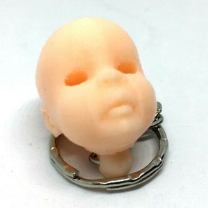 May include: A small, light pink baby doll head keychain with closed eyes and a small mouth. The head is attached to a silver metal keychain.