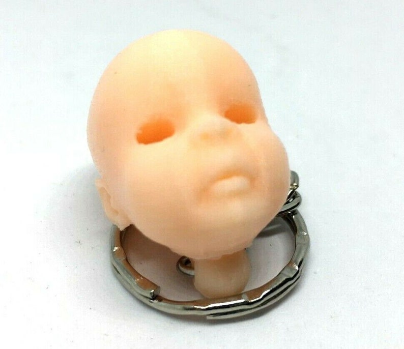May include: A small, light pink baby doll head keychain with closed eyes and a small mouth. The head is attached to a silver metal keychain.