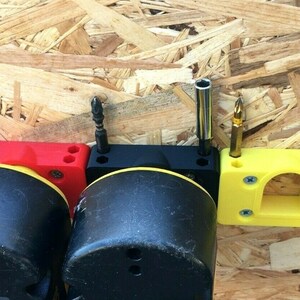 May include: Three 3D printed wall mounts for holding screwdriver bits. The mounts are red, black, and yellow. The mounts have holes for holding the bits and are attached to a wooden surface.