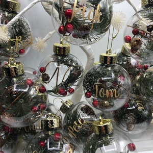 May include: A collection of clear, spherical glass ornaments, each featuring a gold-toned cap and filled with green pine needles, small brown pinecones, and red berries. Each ornament is personalized with a name in gold cursive script.