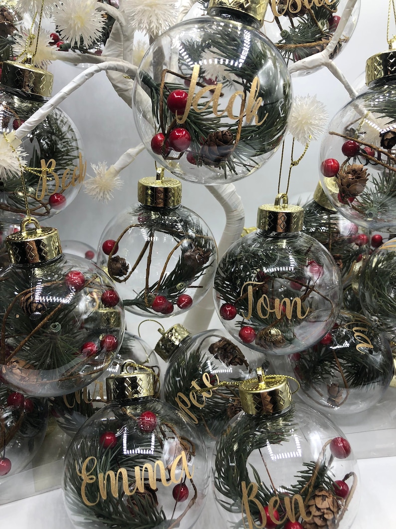 May include: A collection of clear, spherical glass ornaments, each featuring a gold-toned cap and filled with green pine needles, small brown pinecones, and red berries. Each ornament is personalized with a name in gold cursive script.