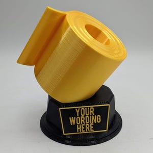 May include: A gold-colored toilet paper roll trophy with a black base. The base has a black plaque with the text "YOUR WORDING HERE".