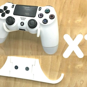 May include: A white PlayStation 4 controller and a matching wall mount are shown against a light wood background. The controller has black buttons and directional controls. The mount is designed to hold the controller securely.
