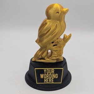 May include: A gold-colored bird figurine perched on a branch, mounted on a black base. The base has a gold-bordered plaque with the text "YOUR WORDING HERE". The bird is detailed with visible feathers.