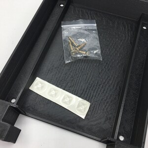 May include: A black plastic box with a textured surface. The box includes four small white adhesive pads and a bag of gold screws.