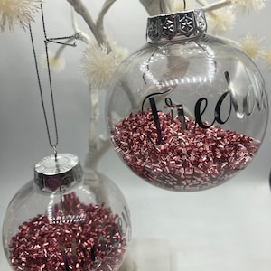 May include: Two clear spherical glass ornaments with silver caps and hanging strings. Each ornament is filled with small, metallic rose gold beads and features a name written in black script: Freddie on the larger one and Lacey on the smaller one.