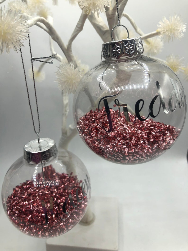 May include: Two clear spherical glass ornaments with silver caps and hanging strings. Each ornament is filled with small, metallic rose gold beads and features a name written in black script: Freddie on the larger one and Lacey on the smaller one.