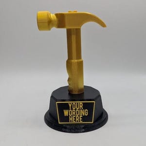 May include: A gold-colored hammer trophy mounted on a black octagonal base. The base features a rectangular plaque with the text "YOUR WORDING HERE" in gold lettering. The hammer has a textured finish and is positioned vertically on the stand.
