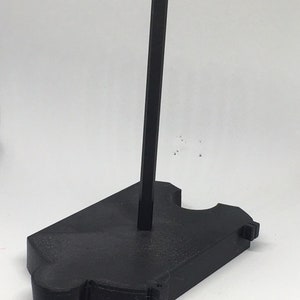 May include: A black plastic display stand with a single vertical rod. The stand has a unique shape with a rounded base and a cutout for holding an object.