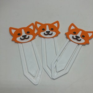 May include: Three white plastic bookmarks with orange and black cartoon dog faces. The dog faces have white eyes and black noses and mouths.