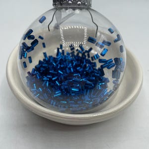 May include: A clear, spherical glass ornament filled with small, metallic blue cylindrical beads. The ornament features a silver-colored metal cap at the top with a white ribbon loop for hanging, resting inside a plain, off-white ceramic bowl.