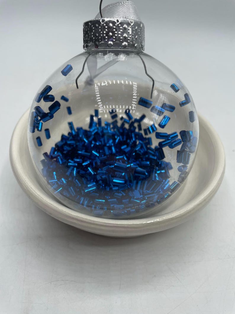 May include: A clear, spherical glass ornament filled with small, metallic blue cylindrical beads. The ornament features a silver-colored metal cap at the top with a white ribbon loop for hanging, resting inside a plain, off-white ceramic bowl.