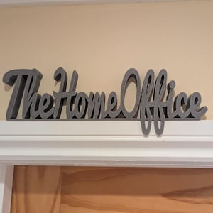 May include: A grey 'The Home Office' sign in a cursive font is mounted above a doorway. The sign is made of a thick material and is in a stylized font. The background is a light beige wall.