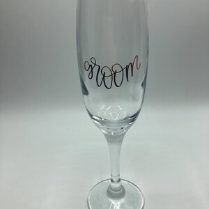 May include: A clear glass champagne flute with the word "groom" written in gold lettering.