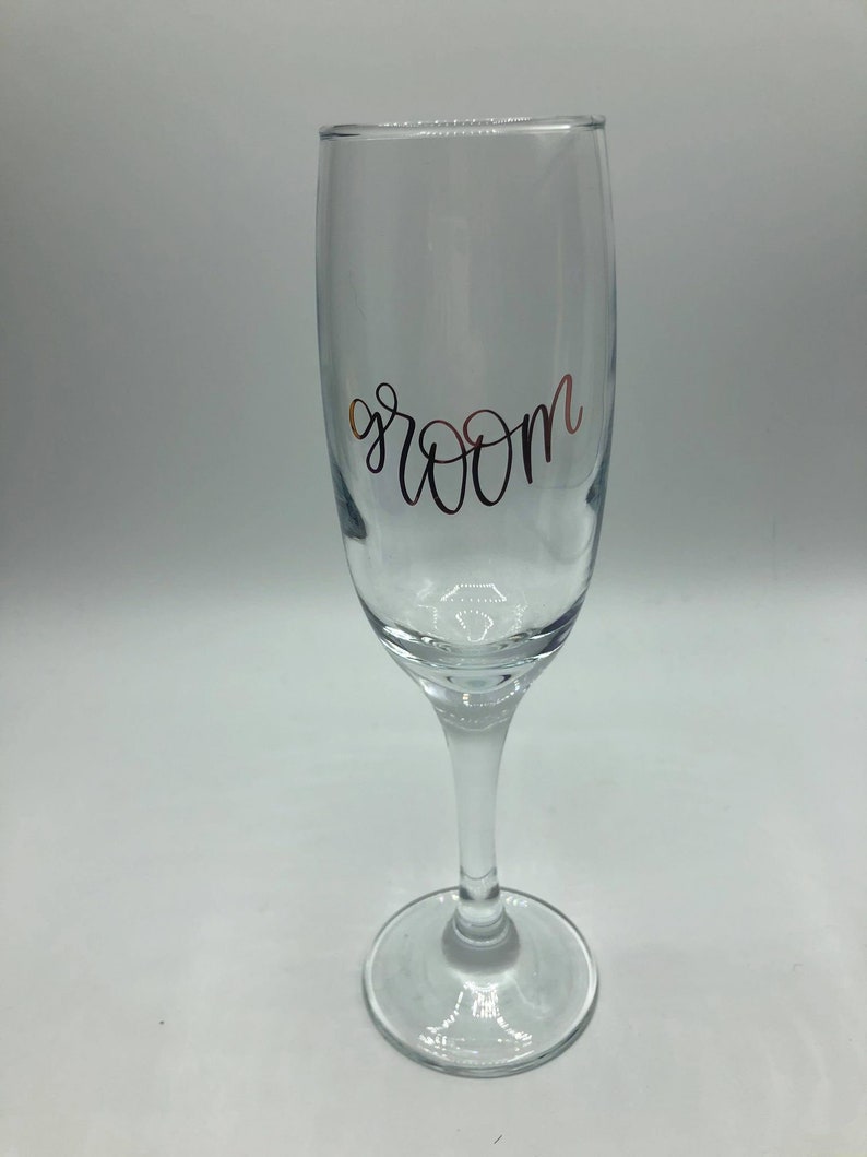 May include: A clear glass champagne flute with the word "groom" written in gold lettering.