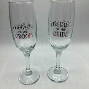 May include: Two clear glass champagne flutes with gold lettering. The left flute says "Mother of the Groom" and the right flute says "Mother of the Bride".