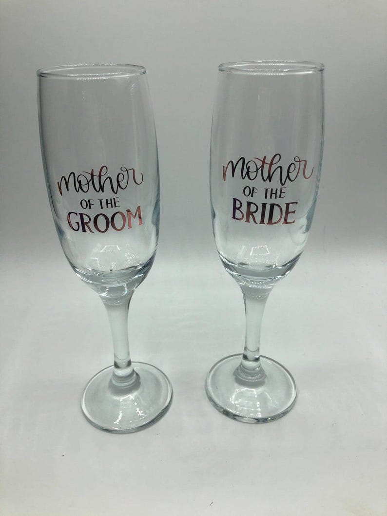 May include: Two clear glass champagne flutes with gold lettering. The left flute says "Mother of the Groom" and the right flute says "Mother of the Bride".