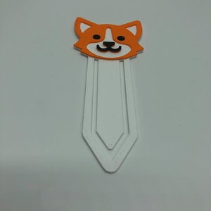 May include: Orange and white bookmark with a cartoon dog face. The bookmark has a white, three-pronged design.