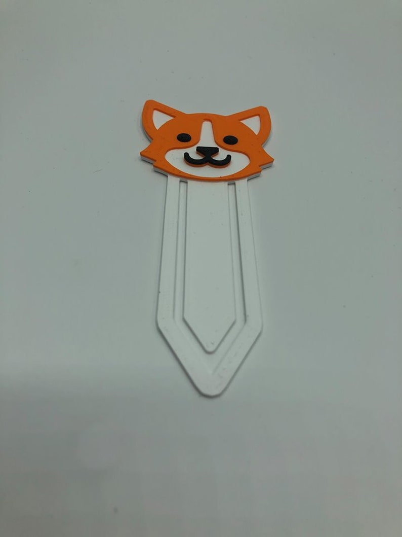 May include: Orange and white bookmark with a cartoon dog face. The bookmark has a white, three-pronged design.