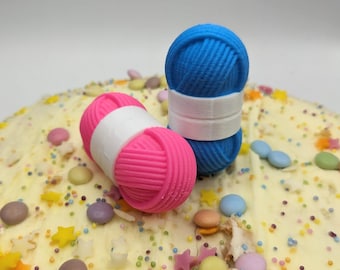 Könnte beinhalten: Zwei Wollknäuel, eines rosa und eines blau, mit weißen Bändern darum, liegen auf einem weißen Kuchen mit bunten Streuseln.