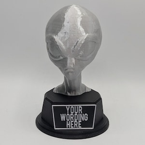 May include: A gray alien head sculpture on a black base with a plaque that says "YOUR WORDING HERE".