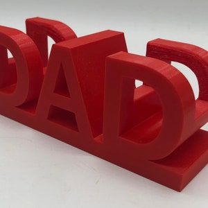 May include: A bright red, three-dimensional plastic sign featuring the word DAD in bold, block capital letters. The letters are connected at the base, creating a sturdy, freestanding structure with a smooth, matte finish and visible print layers.