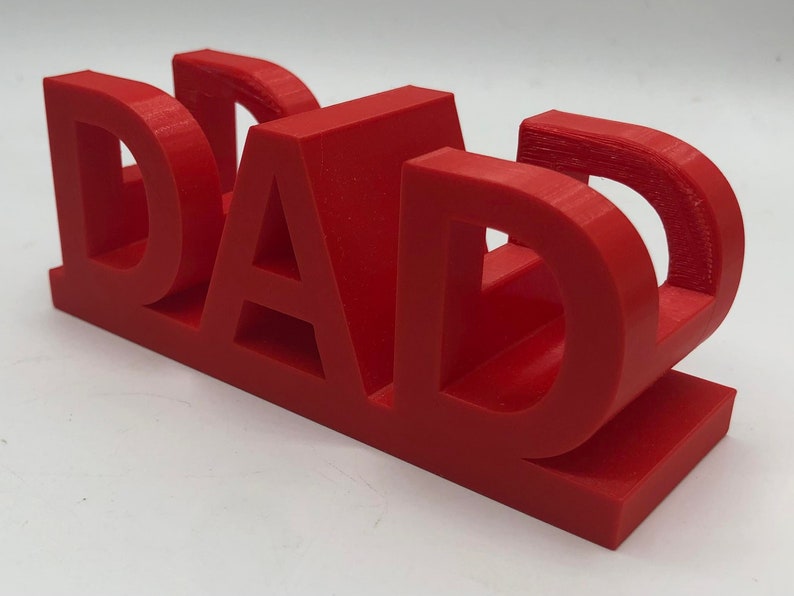 May include: A bright red, three-dimensional plastic sign featuring the word DAD in bold, block capital letters. The letters are connected at the base, creating a sturdy, freestanding structure with a smooth, matte finish and visible print layers.